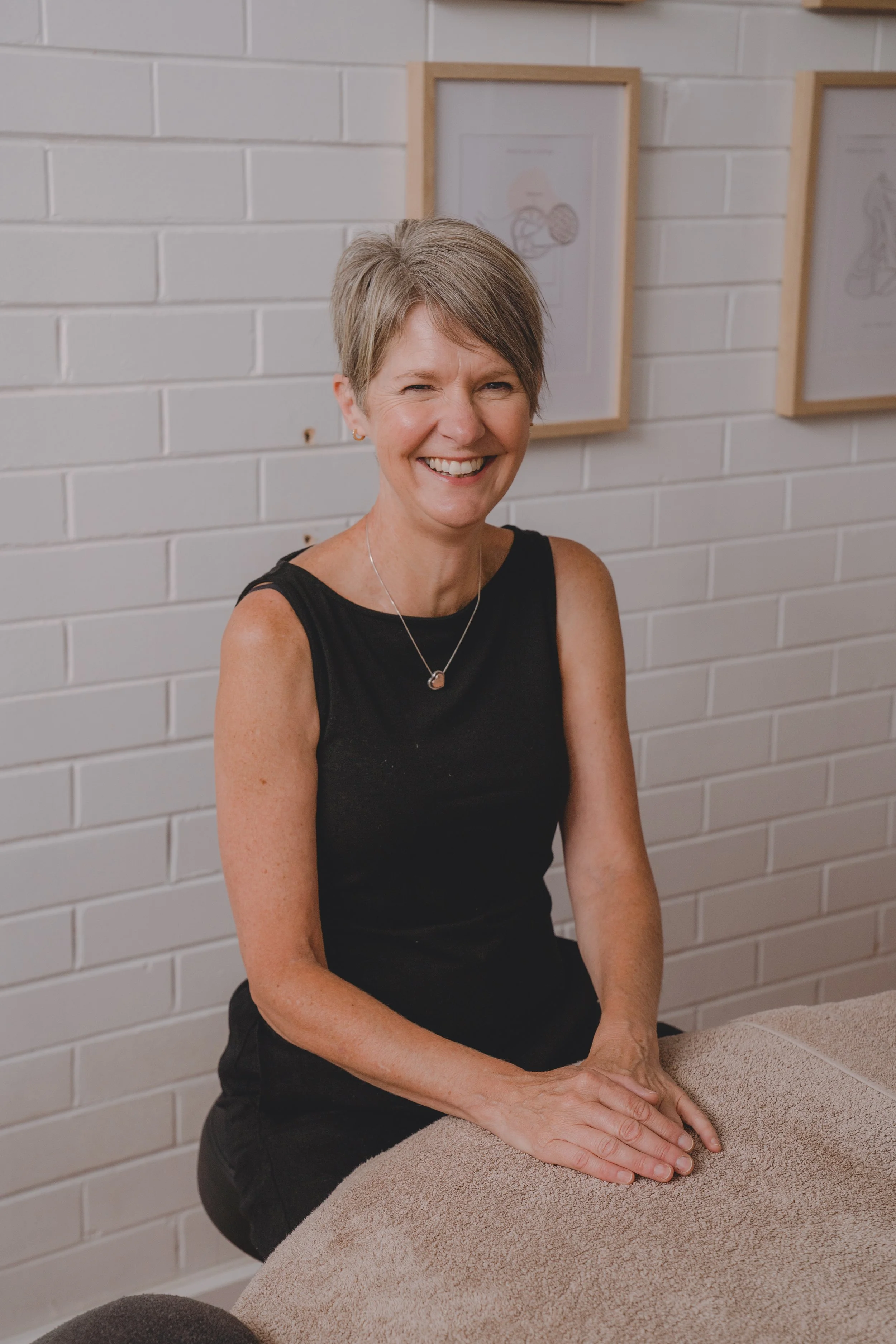 A smiling woman with short gray hair, wearing a black sleeveless top, sitting at a massage table in a spa or wellness center with white brick walls and framed art in the background.