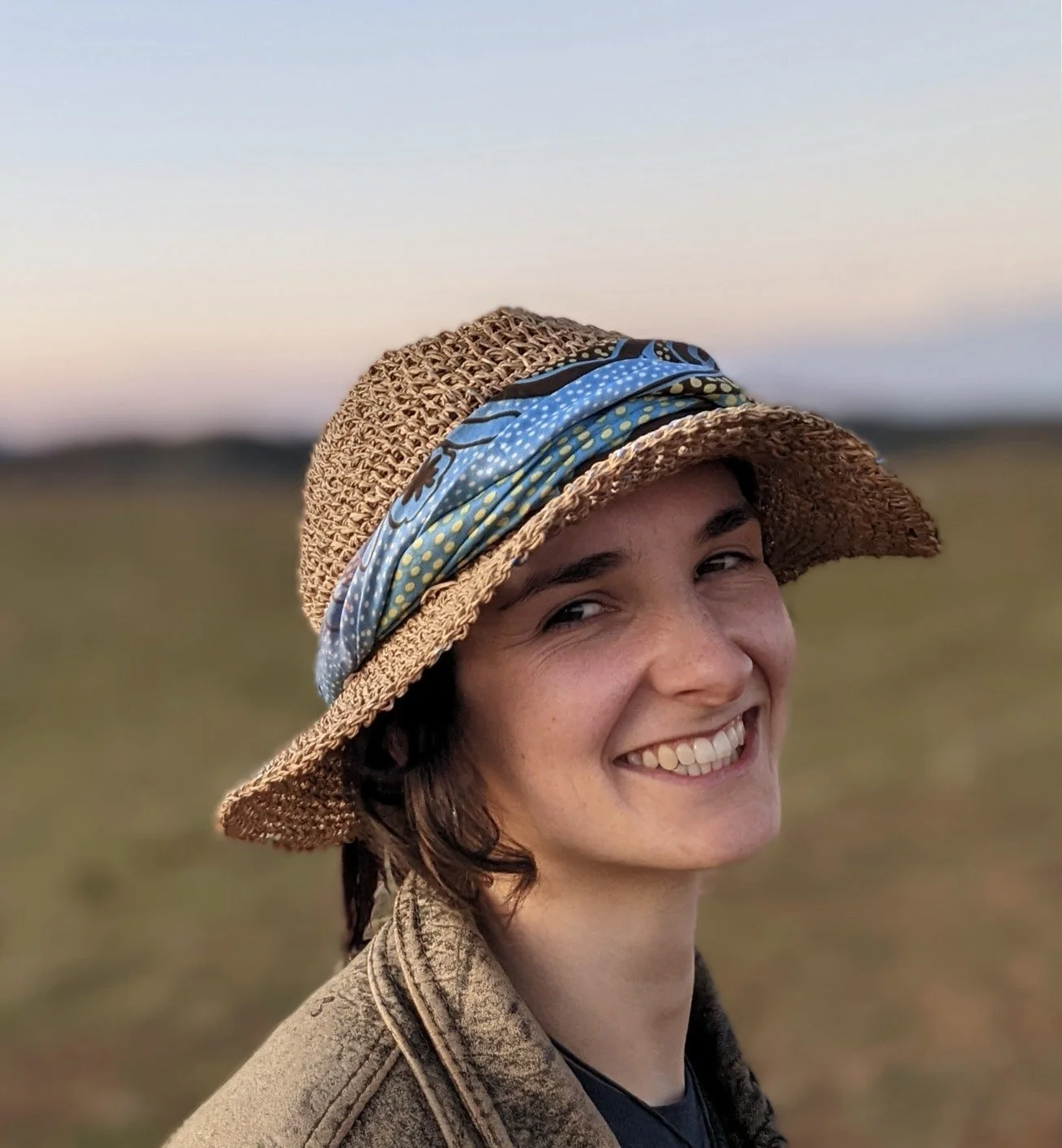 A smiling woman wearing a wide-brimmed straw hat with a blue patterned scarf band, outdoors during sunset with a blurred landscape in the background.