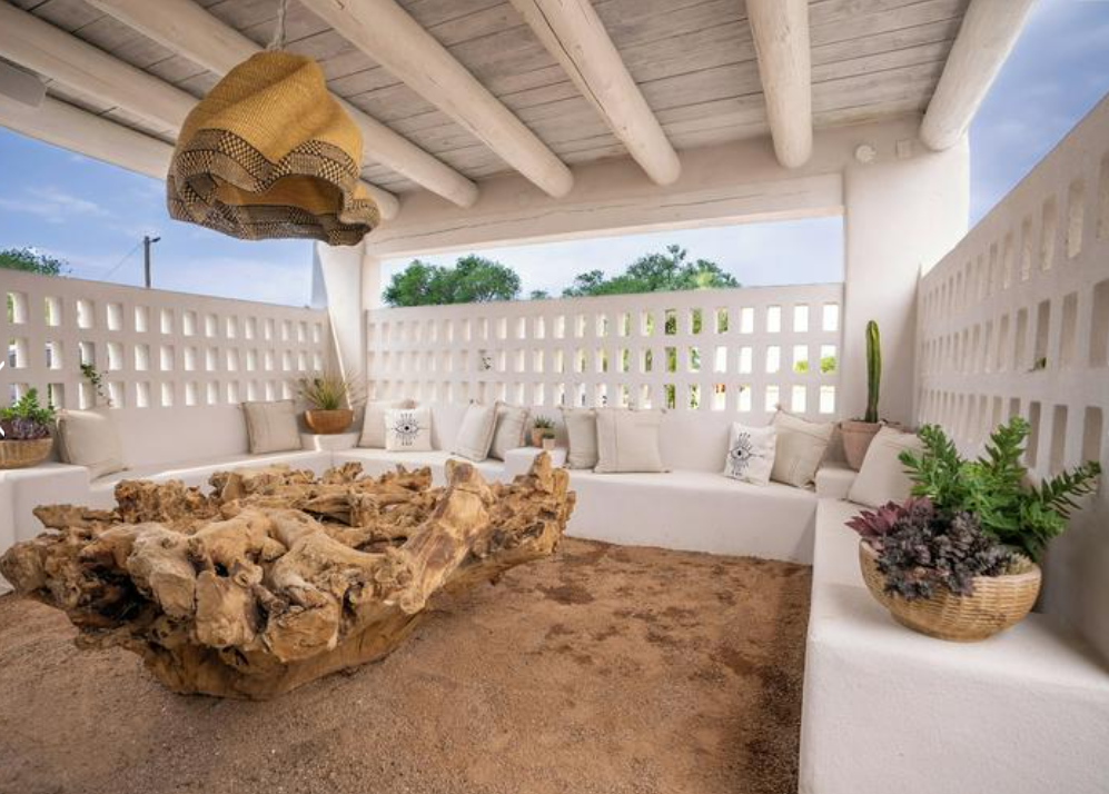 Outdoor patio with a white built-in sectional sofa with beige and patterned throw pillows, a large rustic wooden coffee table, potted plants, and a woven hanging lamp, enclosed by a white lattice wall, with a partial view of the sky and trees.