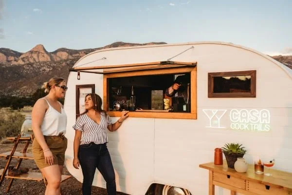 Two women standing outside a small white mobile bar with a sign that says 'Casa Cocktails' in a desert landscape with mountains in the background.