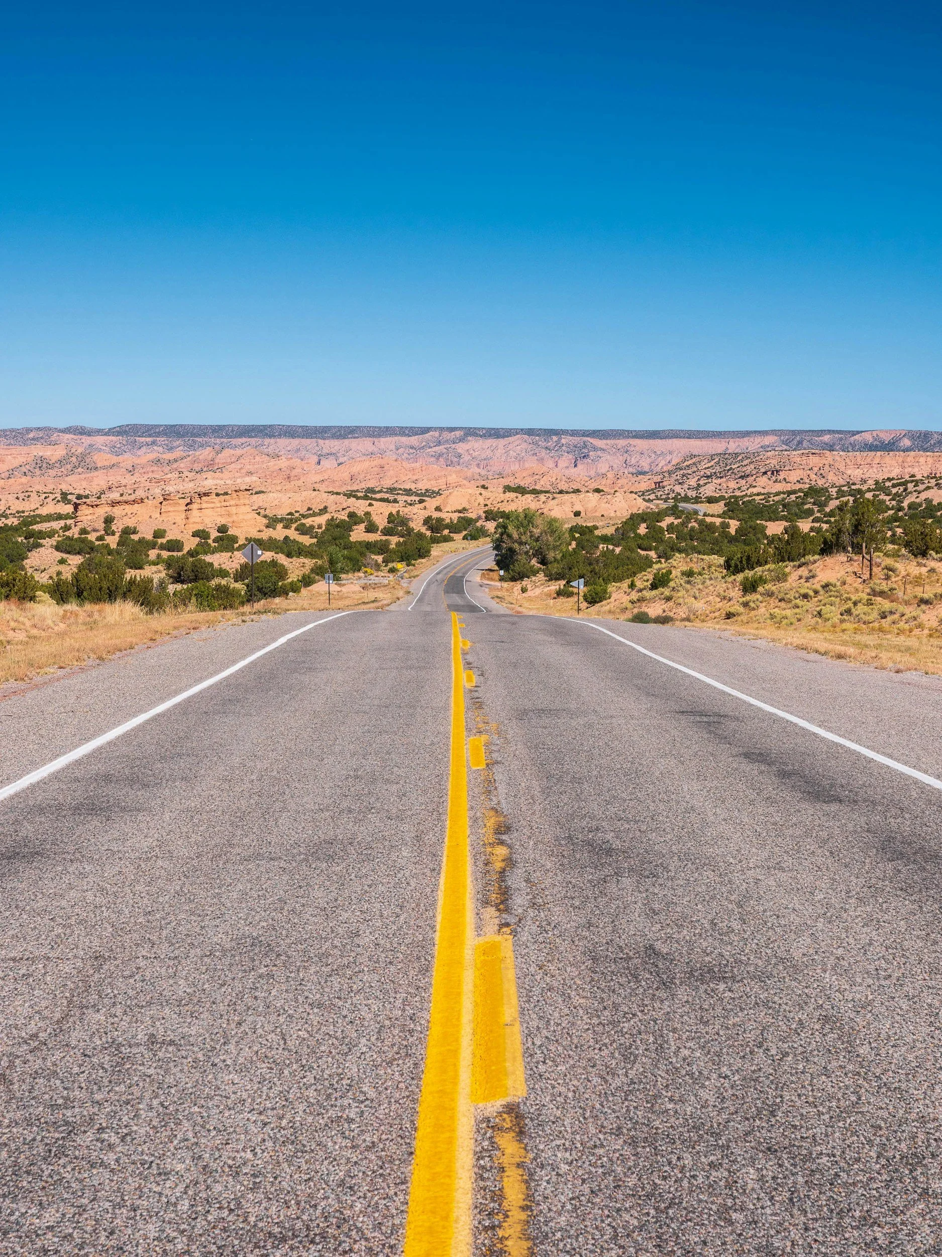 A straight two-lane road stretches into the distance through a desert landscape with rocky hills and sparse vegetation, under a clear blue sky.