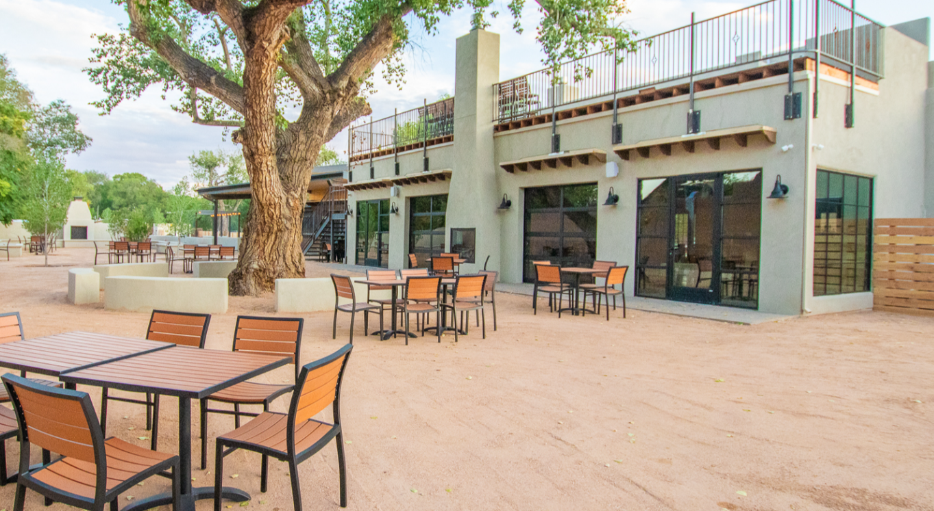 Outdoor patio area with tables and chairs, large tree, and a modern two-story building with large windows and a balcony.