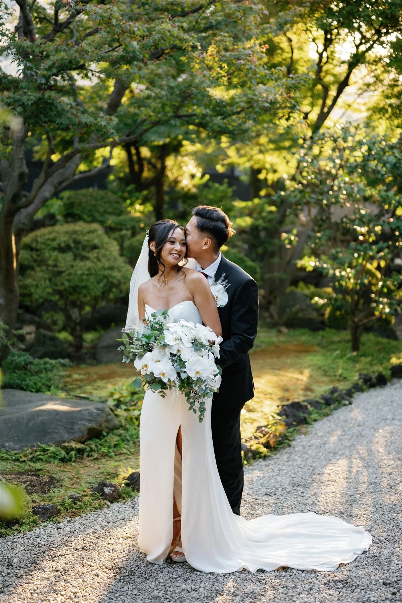 A newlywed couple sharing a kiss outdoors in a lush garden setting, with sunlight filtering through trees, the bride wearing a strapless white gown holding a large bouquet, and the groom in a black suit.