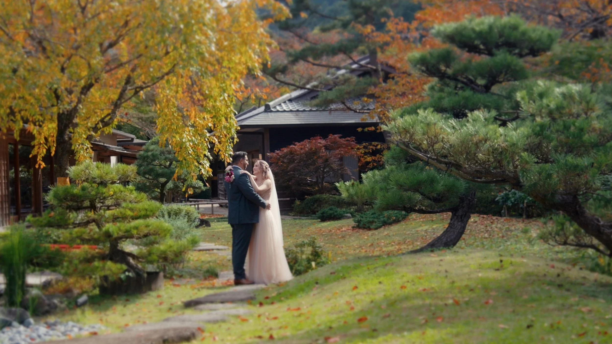 A bride and groom standing close together in a beautifully landscaped garden with autumn foliage, trees, and a traditional building in the background.
