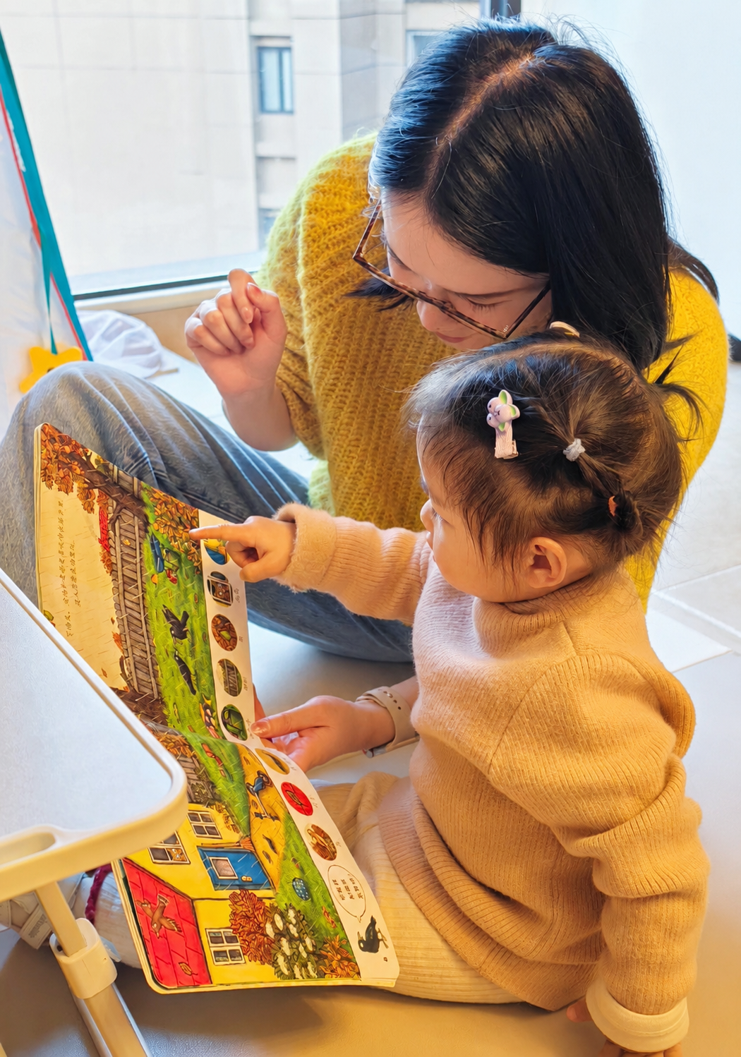 A woman and a young girl sit on the floor reading a colorful children's book with pictures of animals, houses, and train tracks. The girl points at the book while the woman looks on.