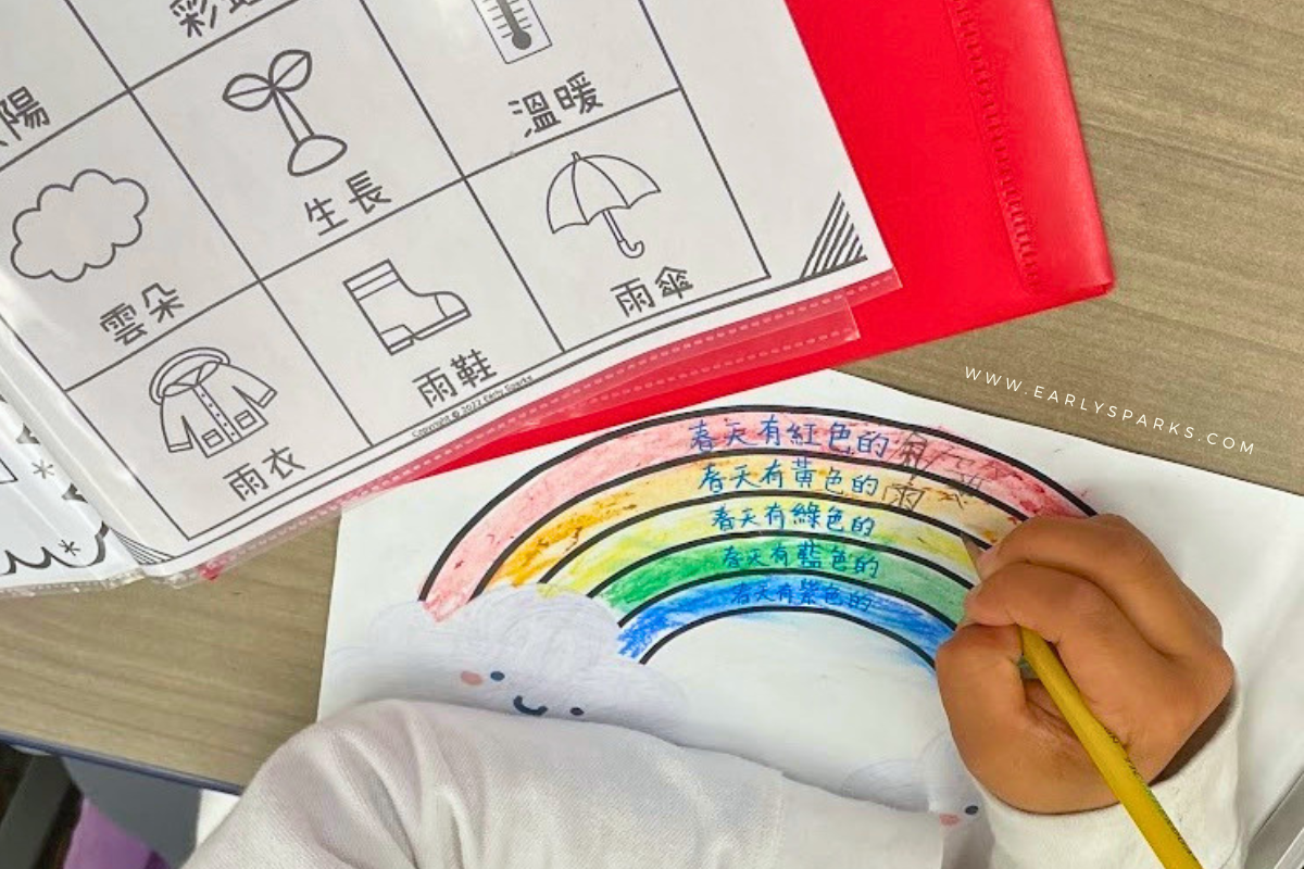 Student writing a Chinese color poem about spring on a rainbow worksheet in a Chinese immersion classroom, using a Mandarin picture dictionary in the folder