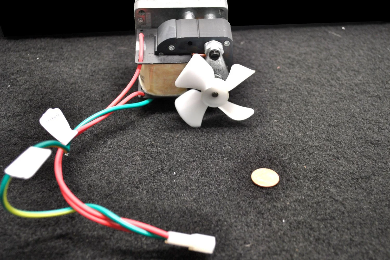 Small electric motor with a white propeller attached, connected to multicolored wires on a black surface, with a penny coin nearby.