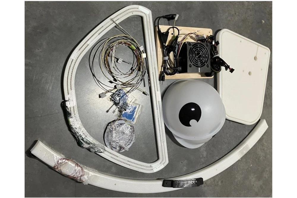 Disassembled electronic components, including a circular LED light frame, wiring, a black electronic module, a white spherical object, and plastic panels, are laid out on a gray surface.
