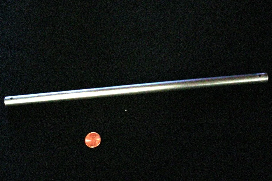 The image shows a long metallic object, possibly a telescoping rod or pipe, floating against a black background, with a penny located below it for scale.