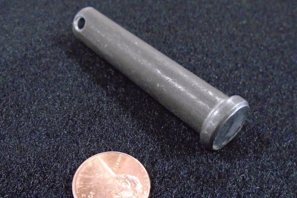 A metal cylindrical pin or rod next to a penny for size comparison on a black textured surface.