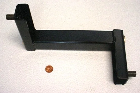 A black L-shaped metal tool with protruding pegs on each end, placed on a white surface next to a penny for scale.