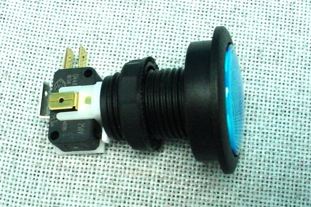 An industrial push button switch with a black focus ring and a blue lens, lying on a dotted fabric surface.