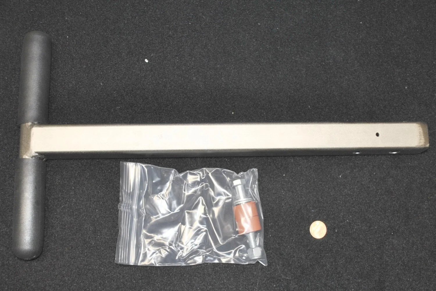 A metal pull-up bar with black foam handles, a small plastic bag containing a screw and a metal bolt, and a penny, all placed on a black textured surface.