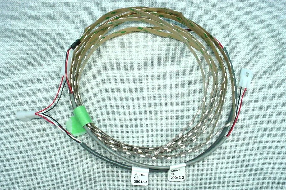 A medical device consisting of multiple flexible metallic strips with attached wiring, labeled with tags reading 'Middle CE 29043-1' and 'Middle CE 29043-2', placed on a light-colored fabric background.