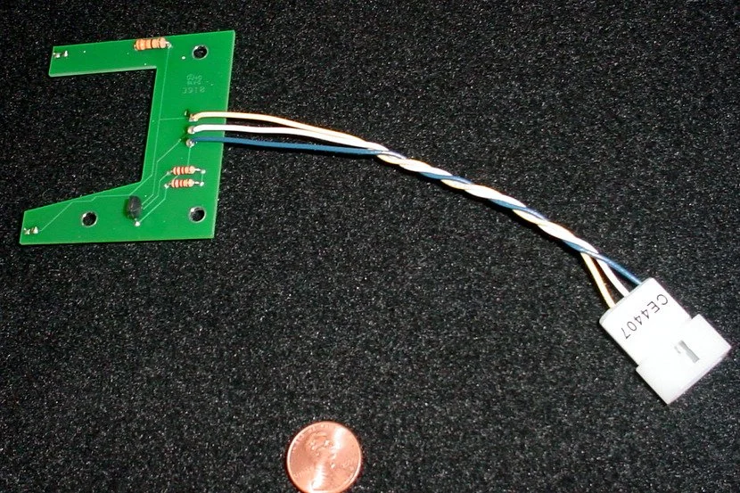 A small electronic circuit board with attached wires and a white connector, placed on a black textured surface next to a U.S. penny for scale.