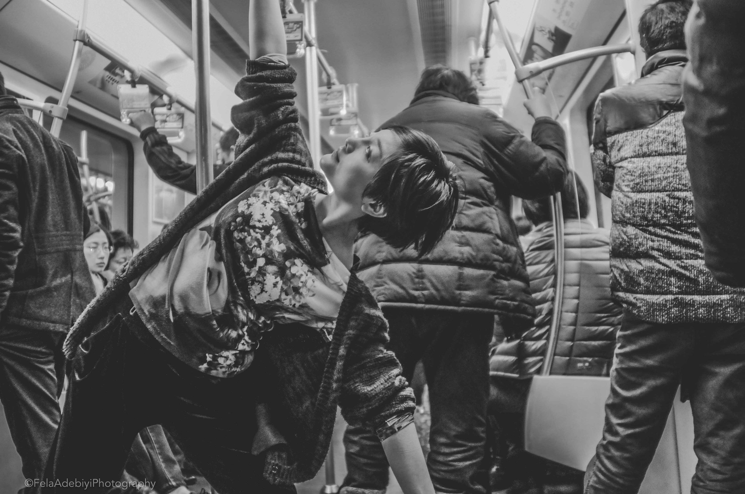 Black and white photo of a person performing a dance pose on a crowded subway, surrounded by standing passengers.