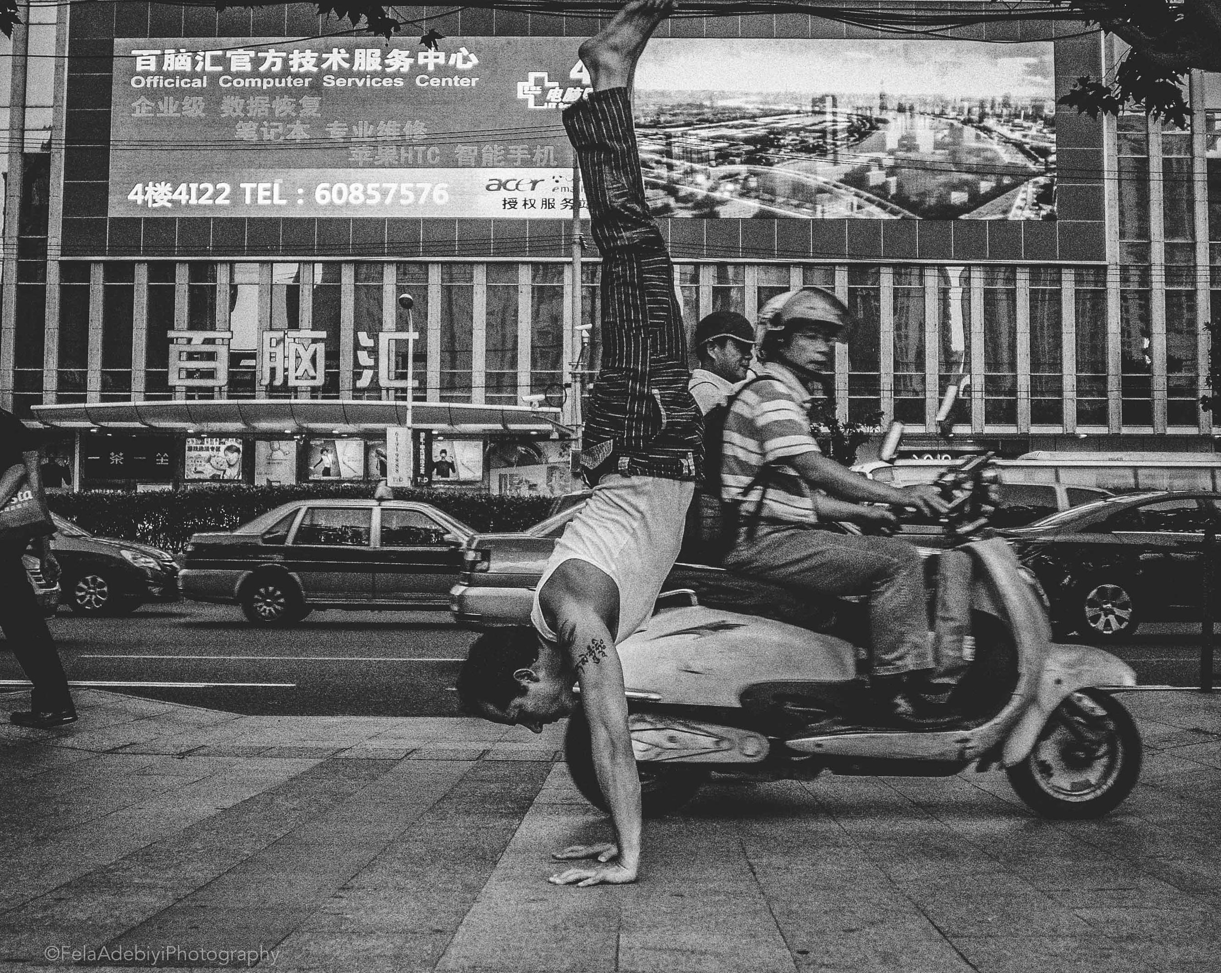 Black and white photo of a man doing a handstand on a sidewalk, with a passing scooter and a busy street background in front of a building with a large sign advertising a computer services center.