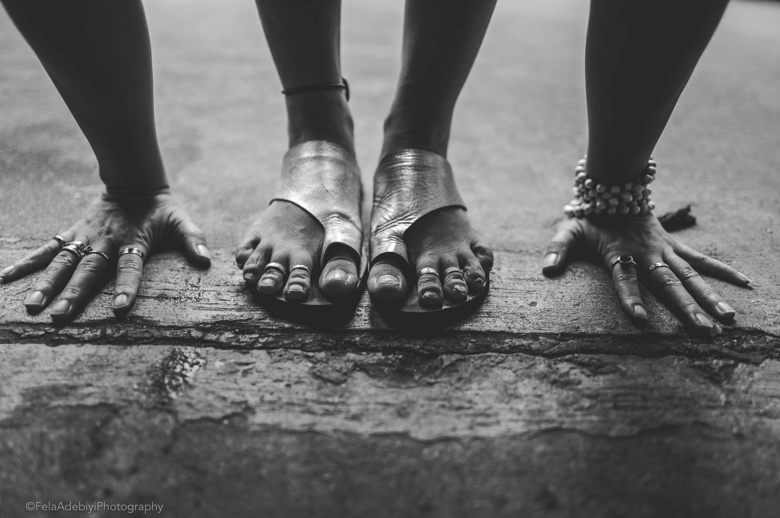 Black and white photo of a person balancing on their hands and feet. Feet are in sandals, and toes have rings. Hands are adorned with rings and bracelets.