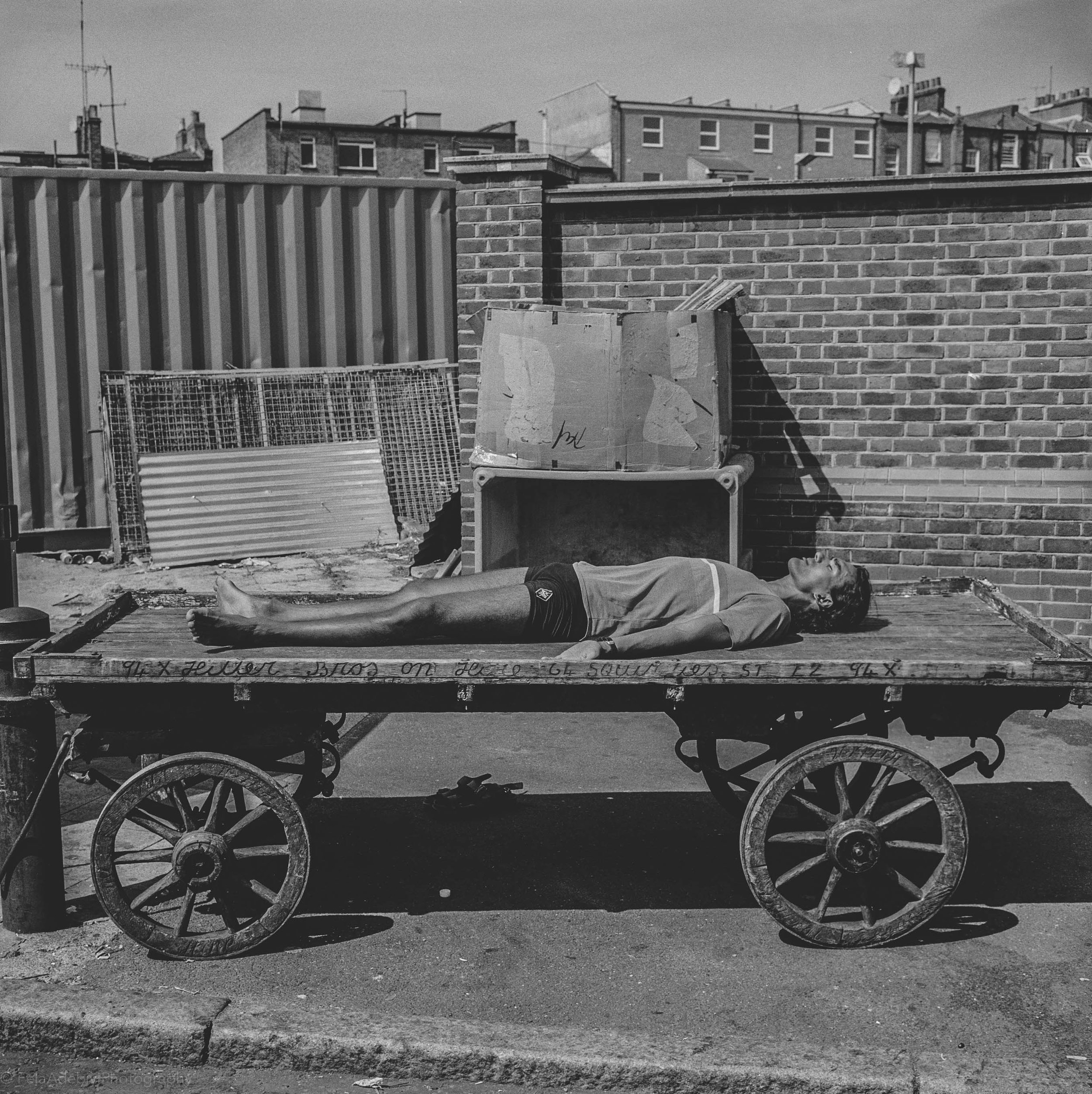 A person is lying on a wooden cart in an urban outdoor setting. The cart is on pavement near a brick wall and surrounded by construction materials and debris.