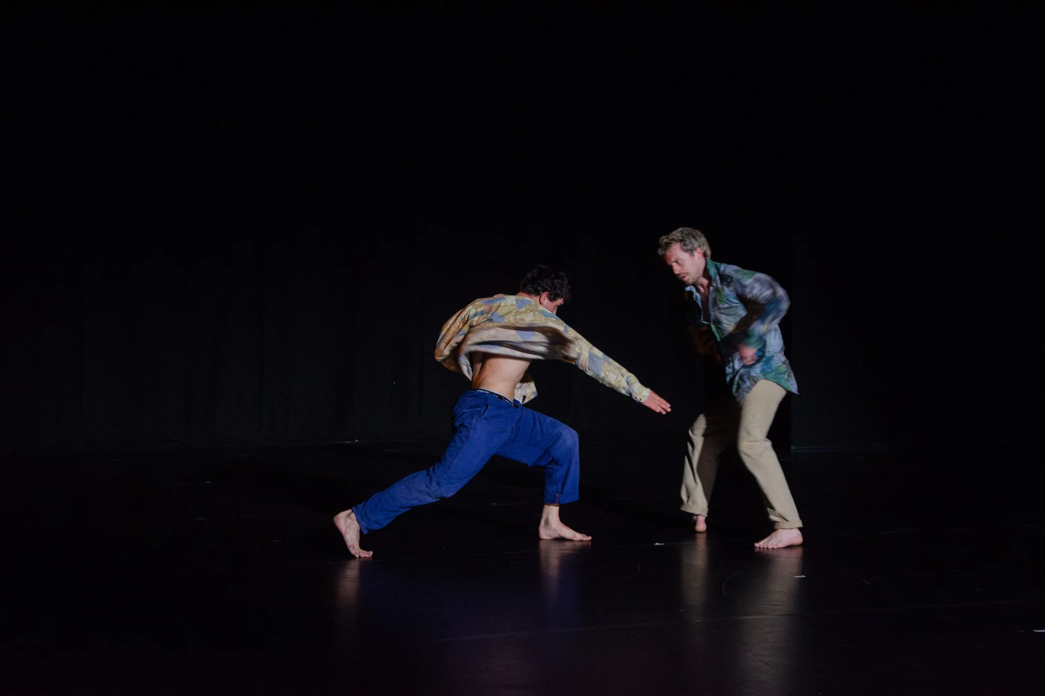 Two men with no shoes practicing dance on a dark stage, one in blue pants and a patterned jacket, the other in beige pants and a patterned shirt, engaged in a dance move.