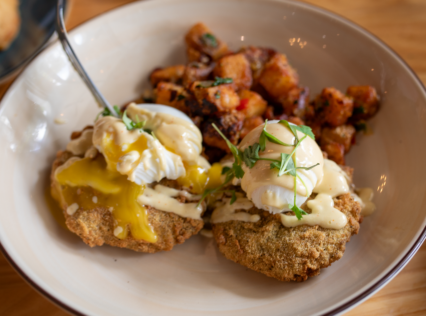 Fried chicken topped with poached eggs, drizzled with hollandaise sauce, served alongside diced, cooked, seasoned potatoes garnished with chopped herbs.