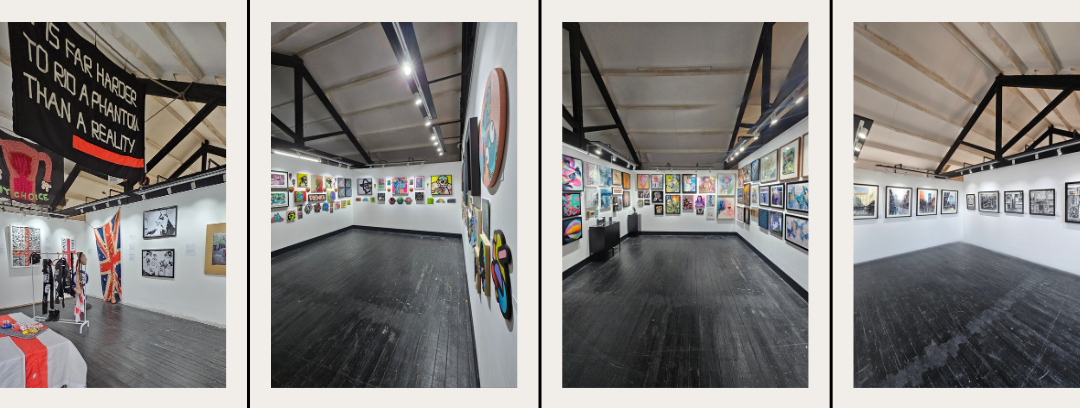 An art gallery with white walls and dark flooring displaying colorful paintings and photographs. There are banners and artwork on the walls, and some sculptures and items on tables.