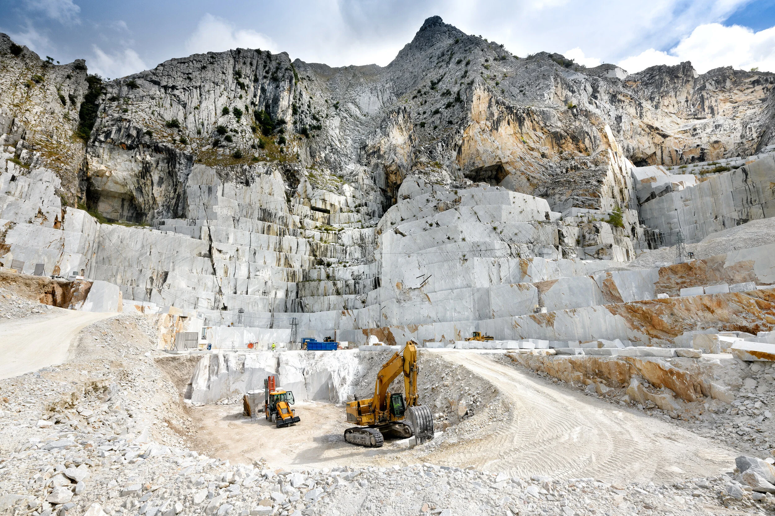 Marble quarry with excavators and raw stone blocks, mountainous backdrop.