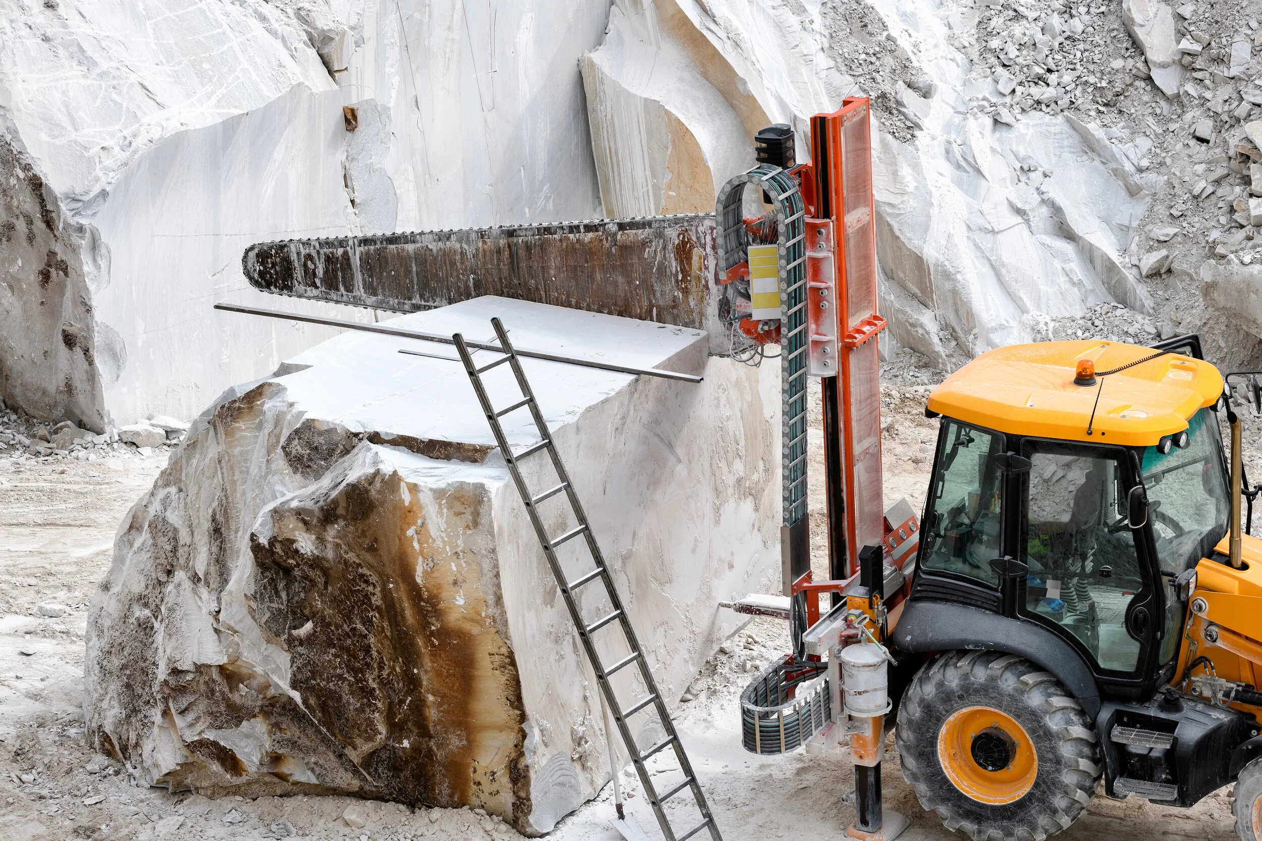 Large industrial saw cutting a marble block with a yellow tractor in a quarry, featuring a ladder leaning on the block.