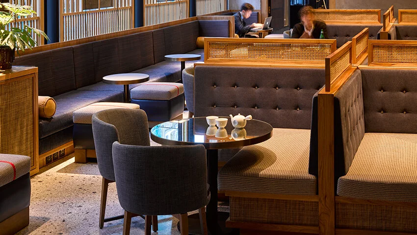 Interior of a stylish cafe with wooden furniture, cushioned seating, tables, and teapots. Two people are seated at separate tables, one using a laptop. A plant is visible on the left side.