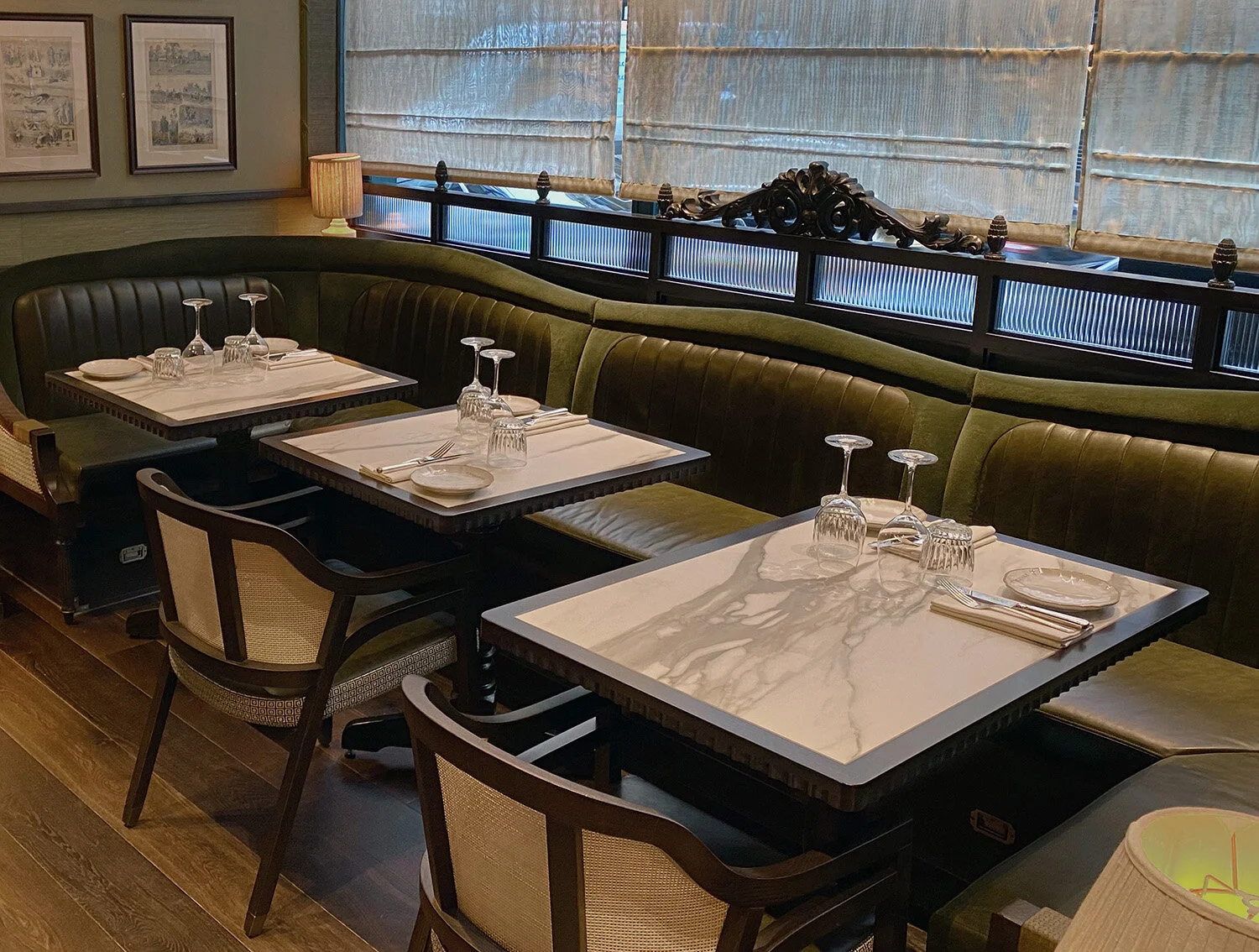 Interior of a stylish restaurant with dark green upholstered banquette seating, marble-topped tables set with glassware and napkins, and framed art on the walls.