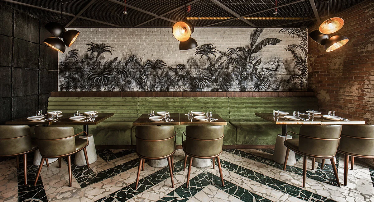 Interior of a modern restaurant with green velvet seating, dining tables set with plates and glasses, patterned tile floor, leafy mural on white brick wall, and contemporary pendant lighting.