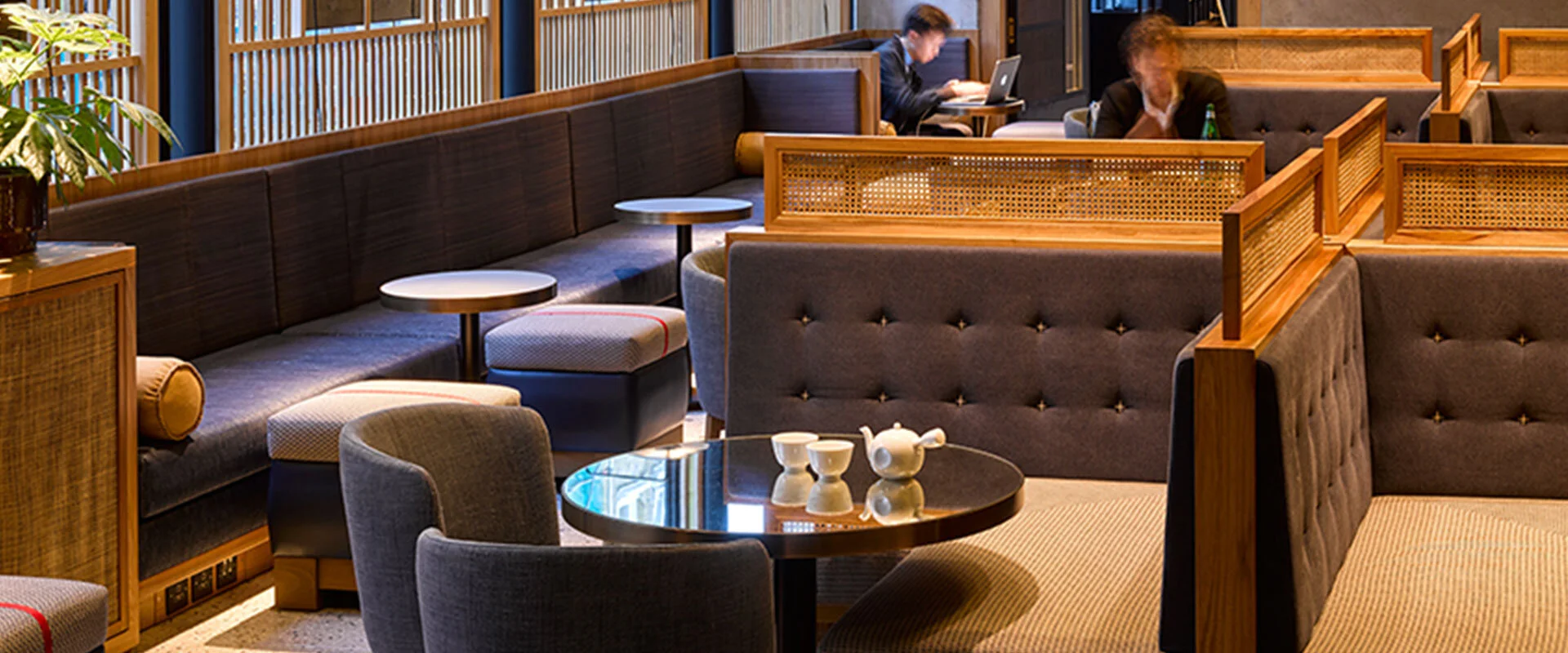 Modern lounge area with cushioned seating, wooden dividers, and small round tables. Two people are working on laptops in the background.