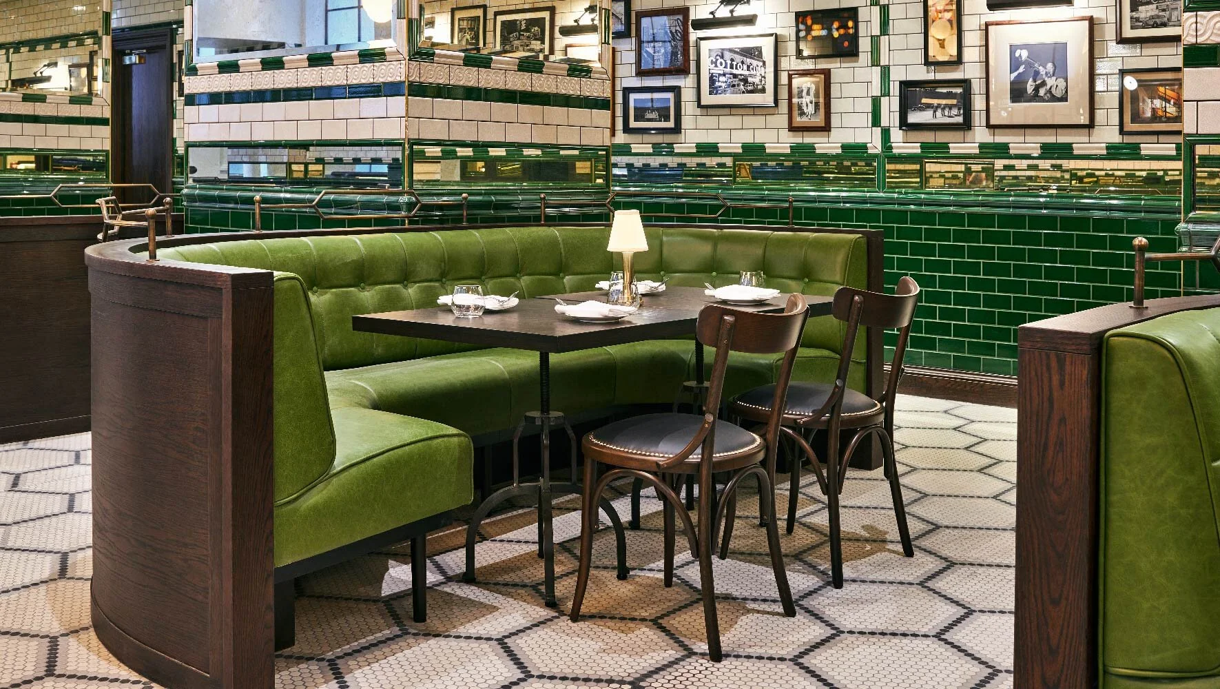 Restaurant interior with green booth seating and dark wood chairs, tiled walls with framed photos, and hexagonal floor tiles.