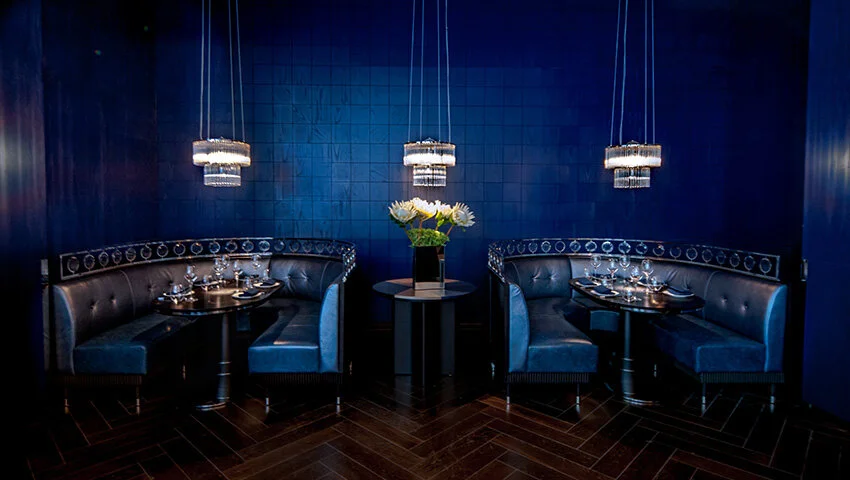 Luxurious blue-themed restaurant interior with curved leather booths, set tables, and elegant hanging chandeliers.