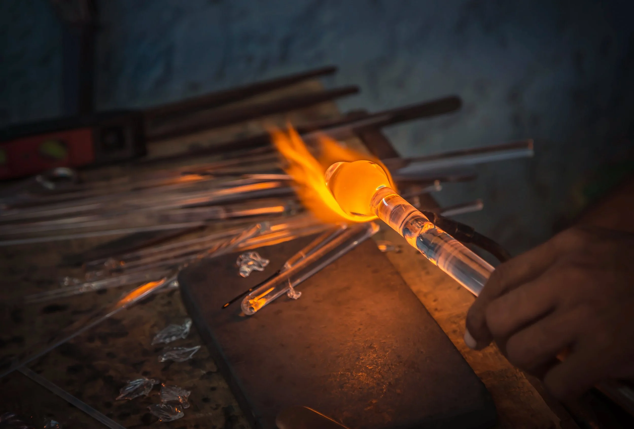 Glassblowing process with person shaping molten glass with tools and flame, surrounded by glass rods on a table.