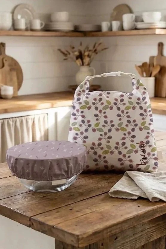 Kitchen with open wooden shelves holding white dishes, bowls, and mugs, some cutting boards, a vase with dried flowers, and a cloth-covered bowl and a fabric bag with a floral pattern on a rustic wooden table.