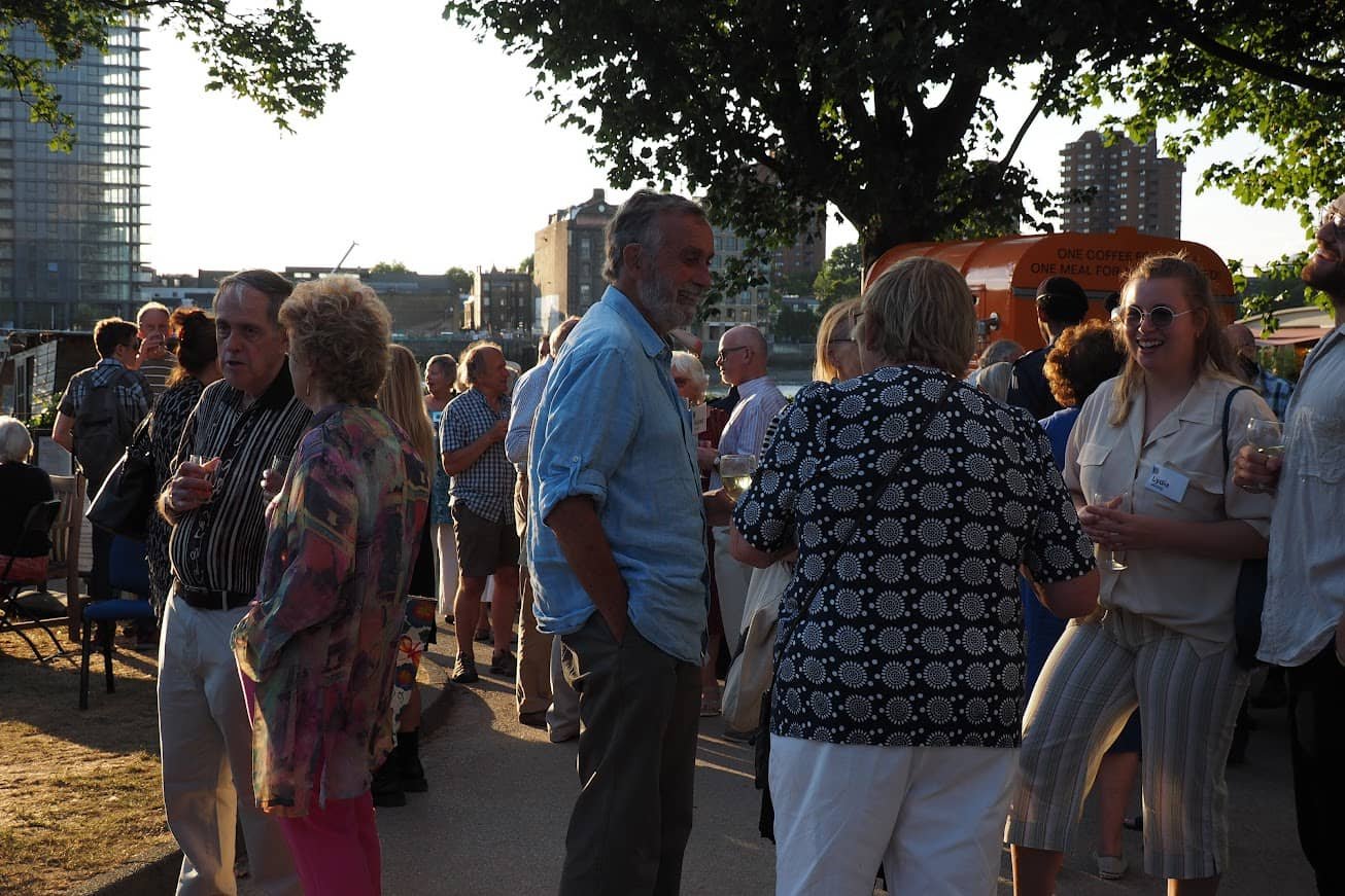 People socializing outdoors near a river with trees and city buildings in the background during sunset.