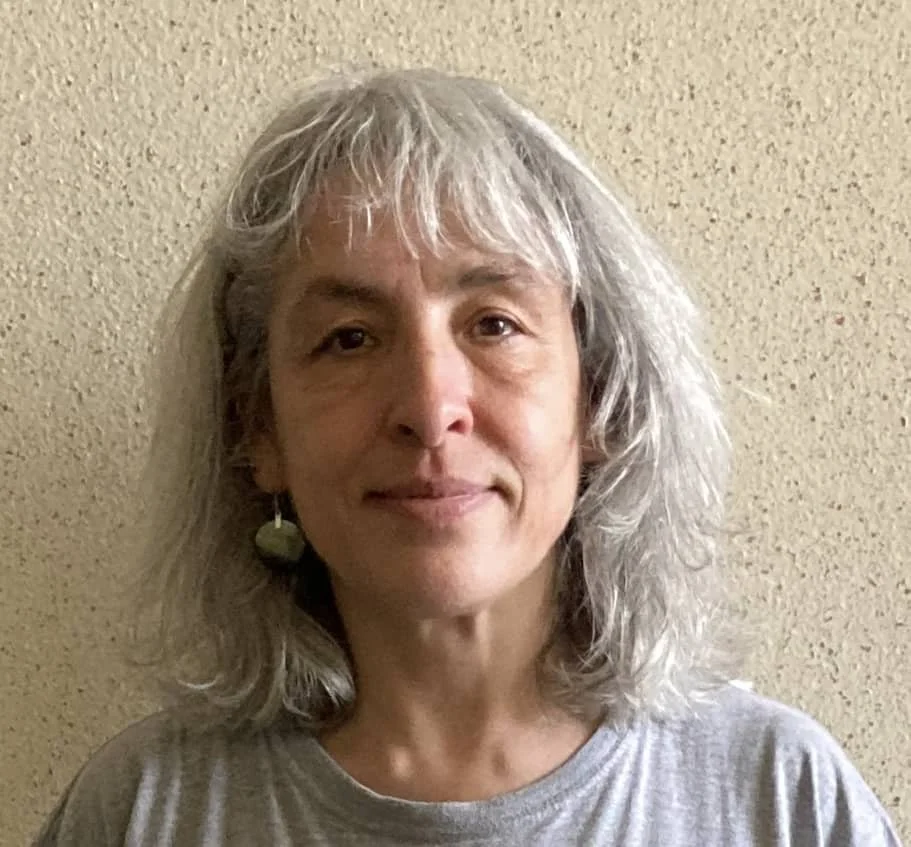 A woman with gray, shoulder-length hair, wearing a light gray shirt and earrings, standing against a beige textured wall.