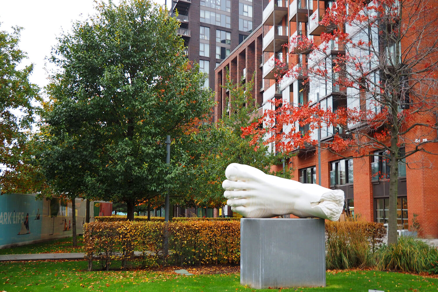 Outdoor city scene with modern multi-story buildings, trees with green and red leaves, and a large white sculpture of a foot on a gray pedestal.