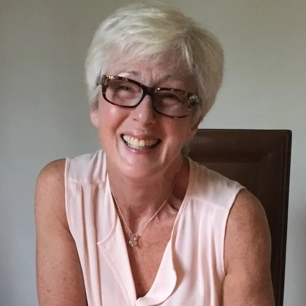Smiling woman with short white hair, wearing glasses and a light pink sleeveless top, sitting in a room with a plain wall behind her.