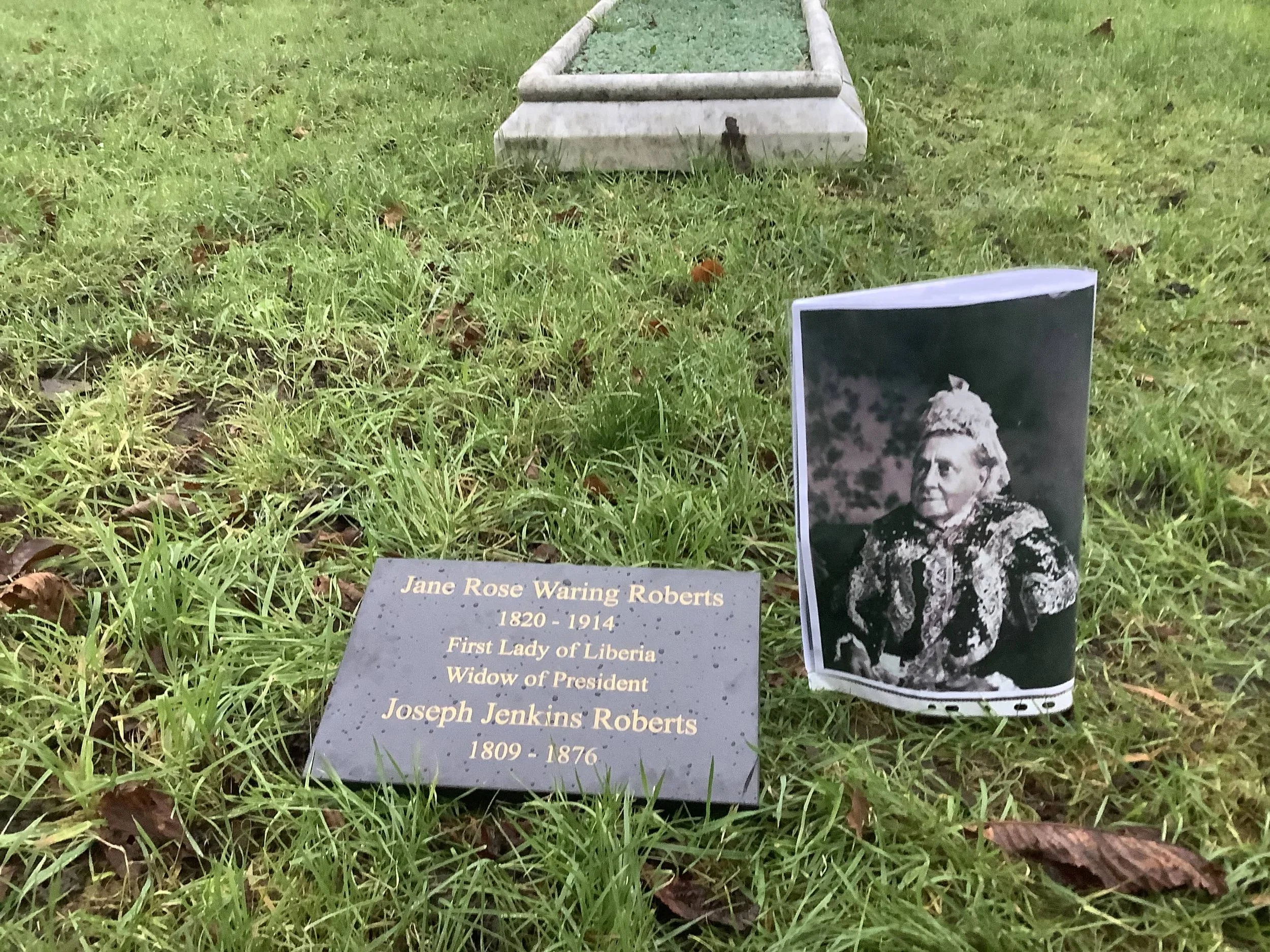 A slate marker engraved with the words: 'Jane Rose Waring Robert 1820-1914 First Lady of Liberia' on grass, with a photo of Jane Roberts next to her and her grave in the background