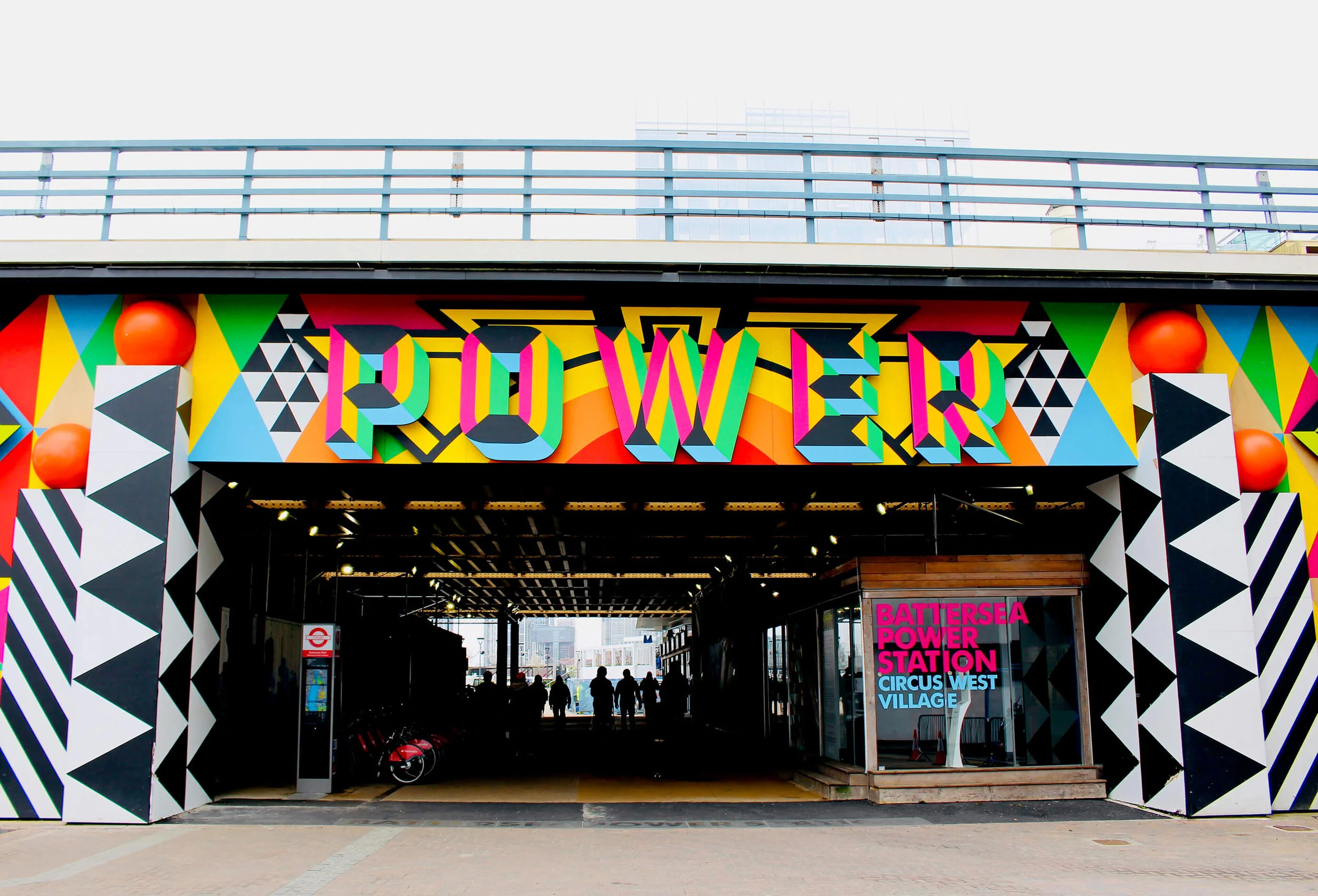 Colorful entrance with the word "POWER" in bright, multicolored 3D letters, geometric patterns, and large orange spheres on the sides, leading into a tunnel with silhouettes of people inside, signage for Battersea Power Station, Circus West Village.