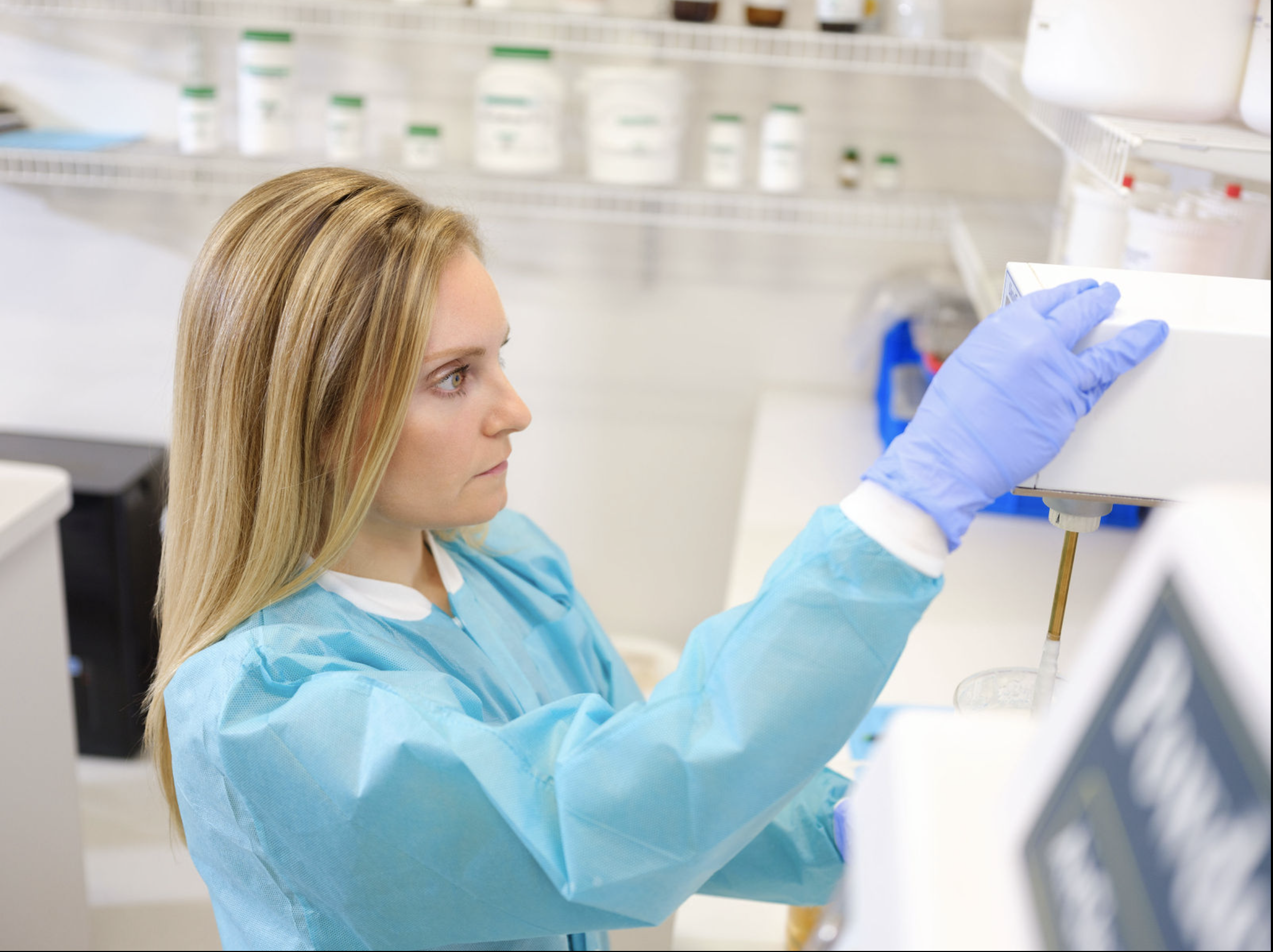 A lab technician in protective gear carefully operates laboratory equipment in a clinical setting surrounded by medical supplies.