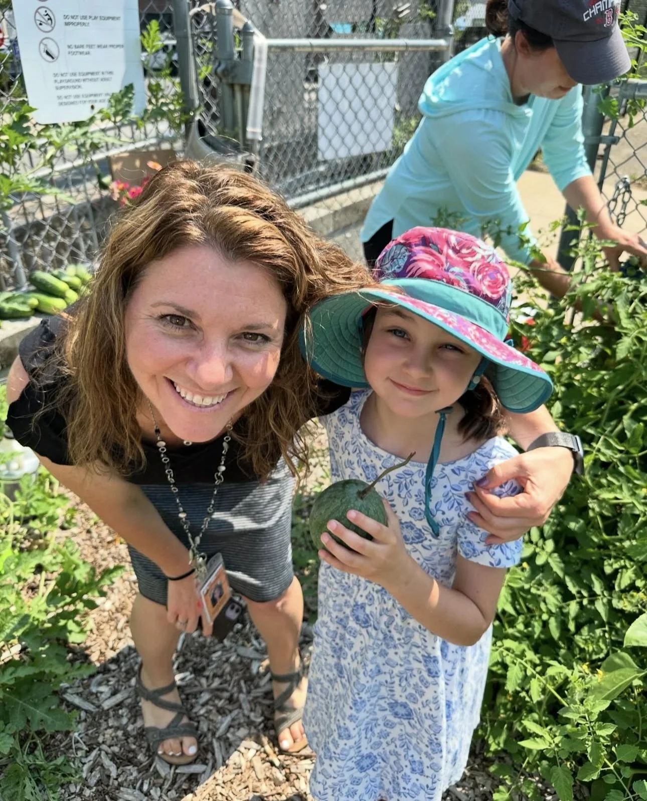 A woman and a young girl smiling outdoors in a garden, holding a small green pumpkin. The girl is wearing a colorful sunhat and a floral dress, while the woman is dressed in casual summer clothing and sandals. In the background, another person is tending to plants near a chain-link fence.