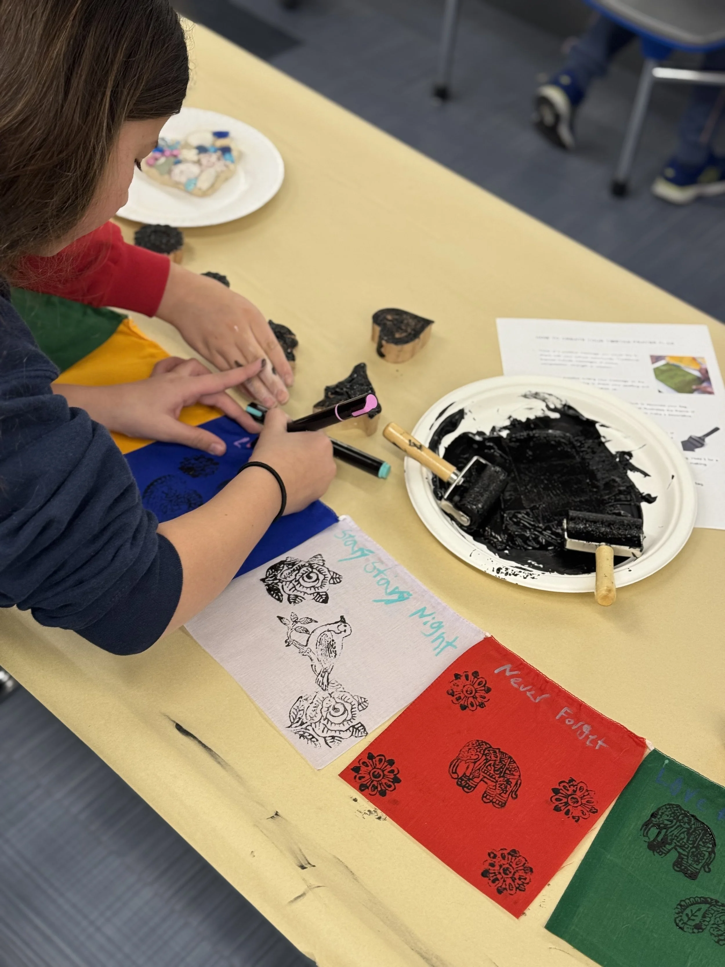 Child carving rubber stamps with black ink on a table, surrounded by printouts and colored papers featuring animal and text designs.