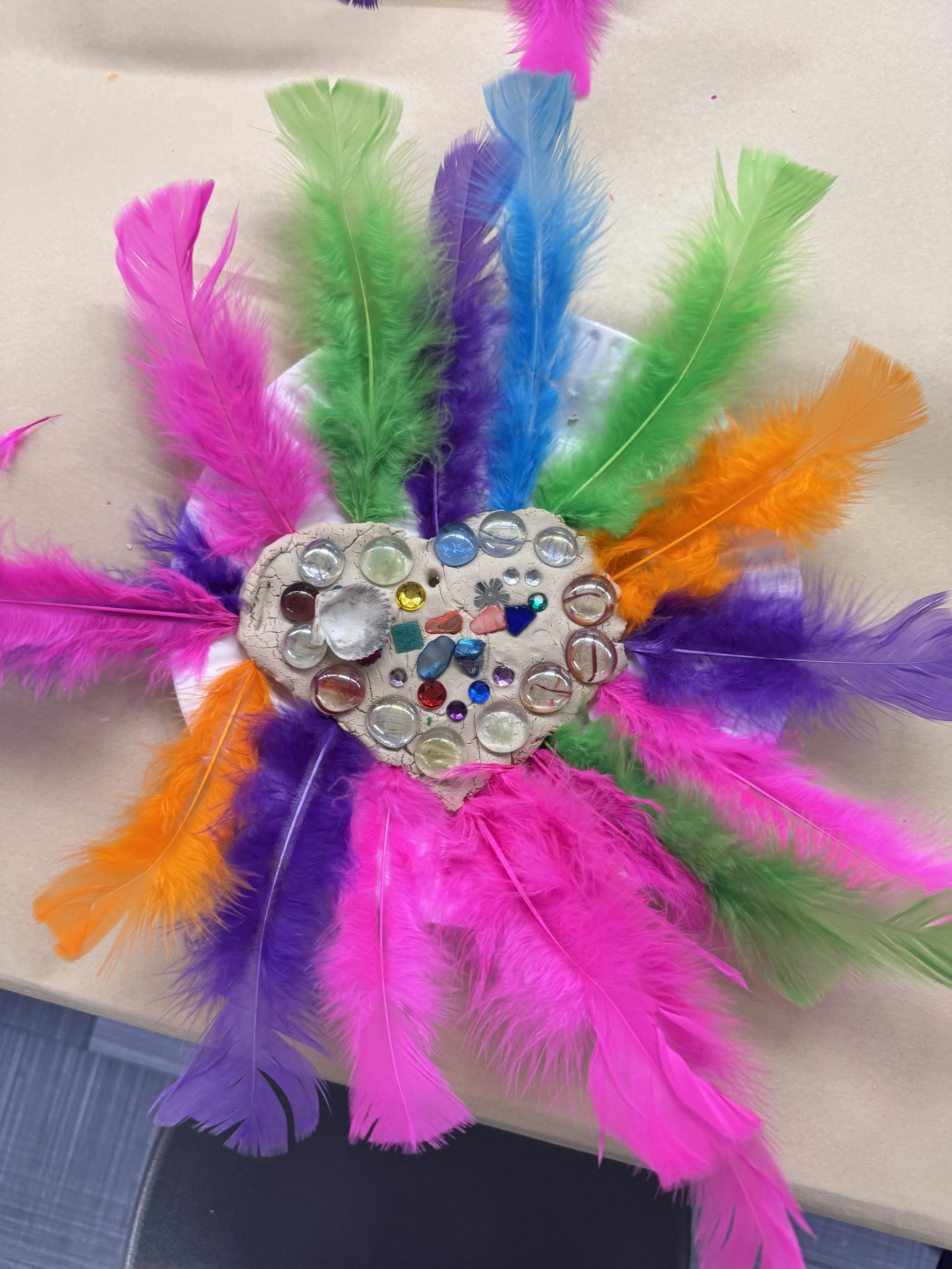 Colorful heart-shaped craft decorated with multicolored feathers, beads, and small decorative objects.