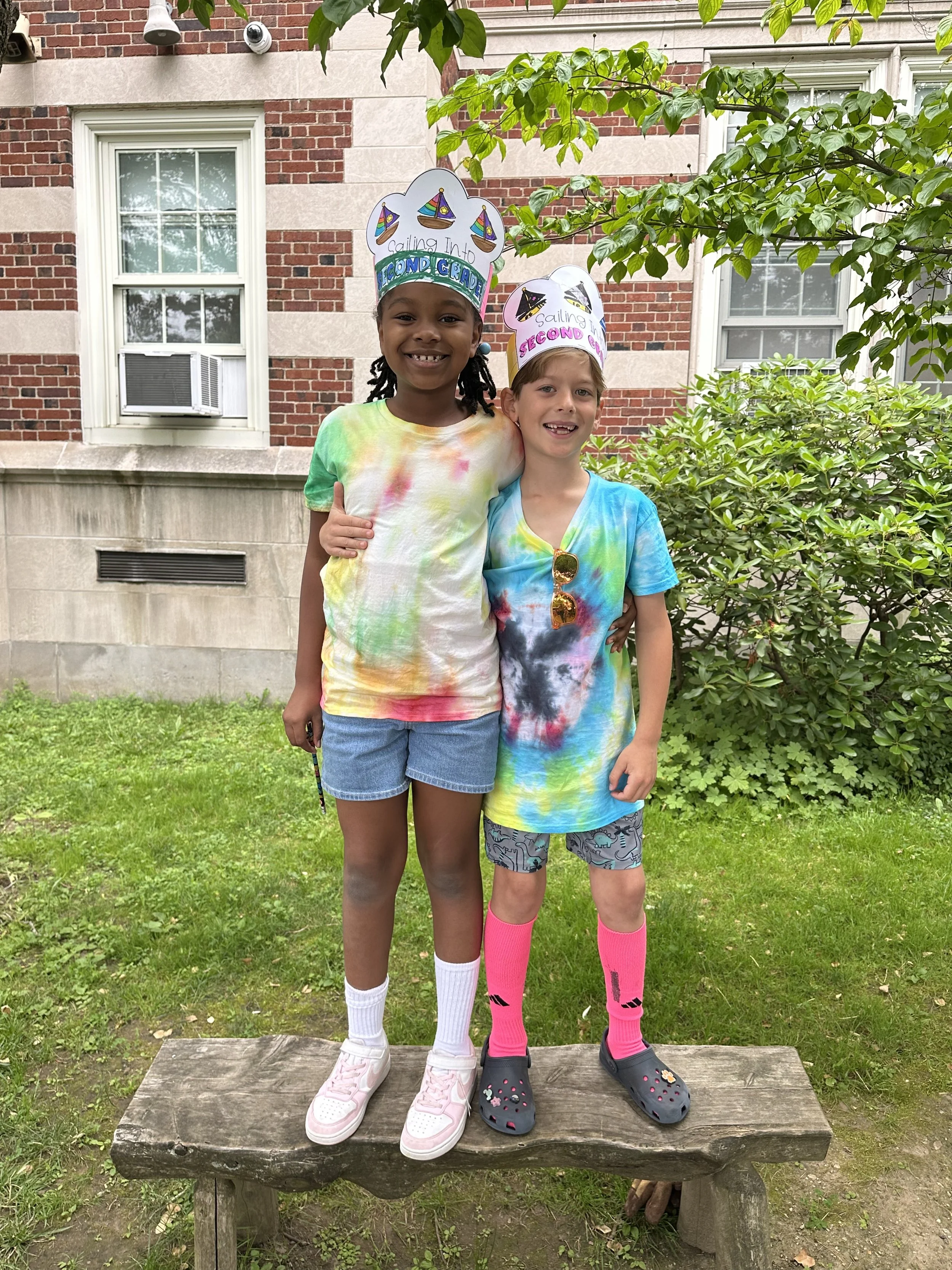Two children standing on a wooden bench outdoors, smiling and wearing colorful tie-dye shirts and paper crowns that say 'Sailing into Second Grade' and 'Sailing into Third Grade.'