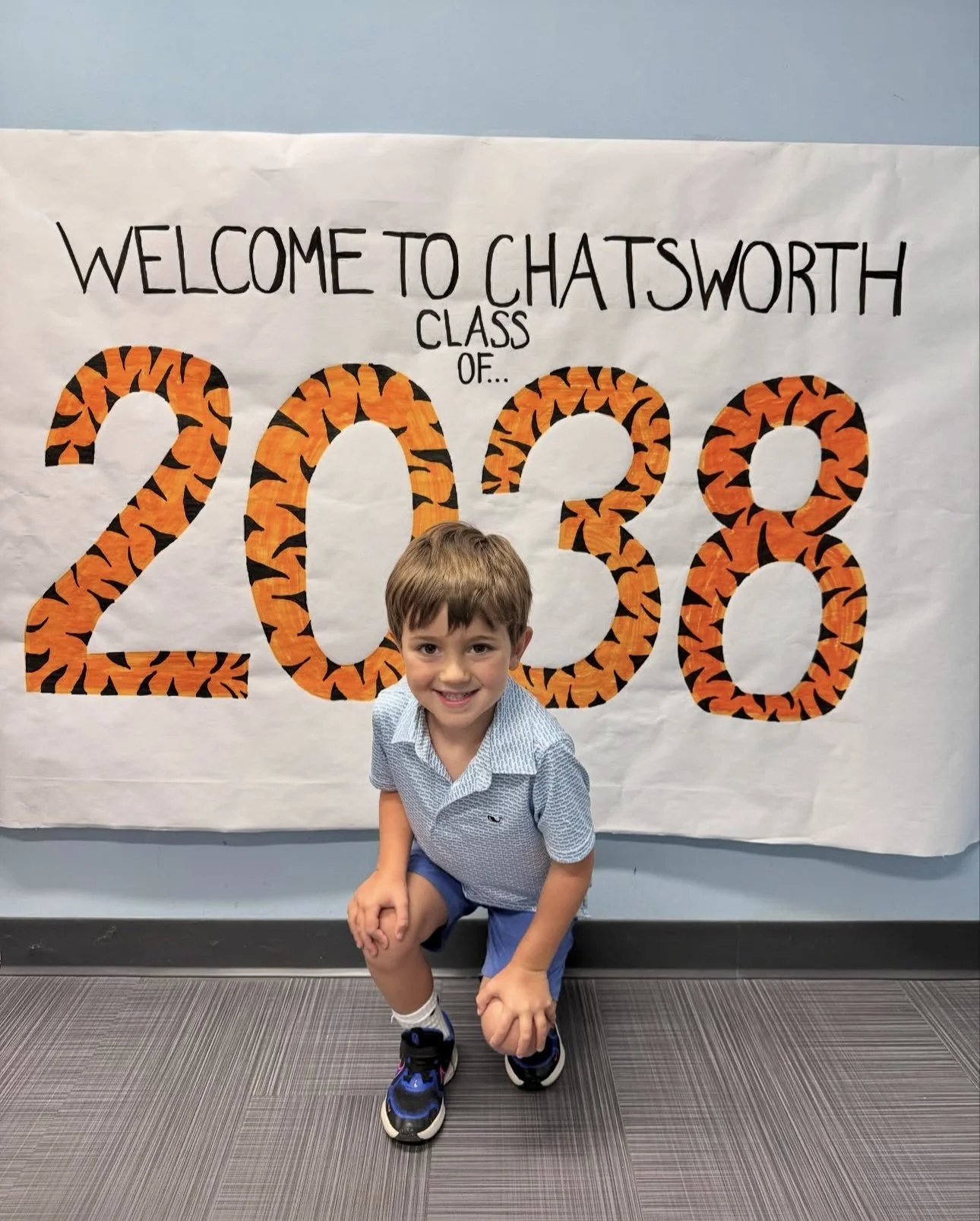 Happy young boy kneeling in front of a large decorated sign for a 2028 class at Chatsworth School, with orange and black tiger-striped numbers.