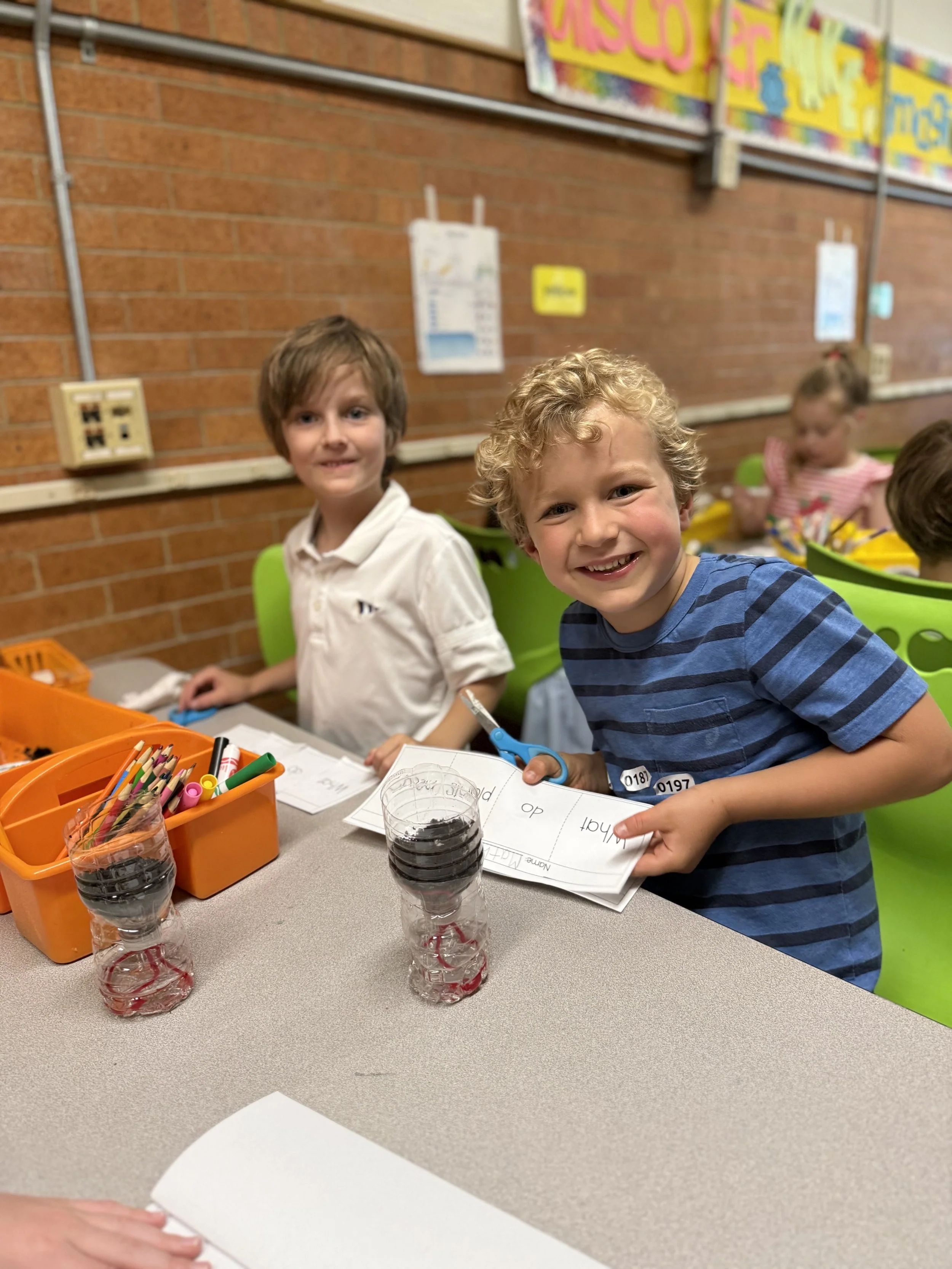 Two young boys in a classroom, smiling and holding scissors, with paper and colored markers on the table. There are other children in the background.