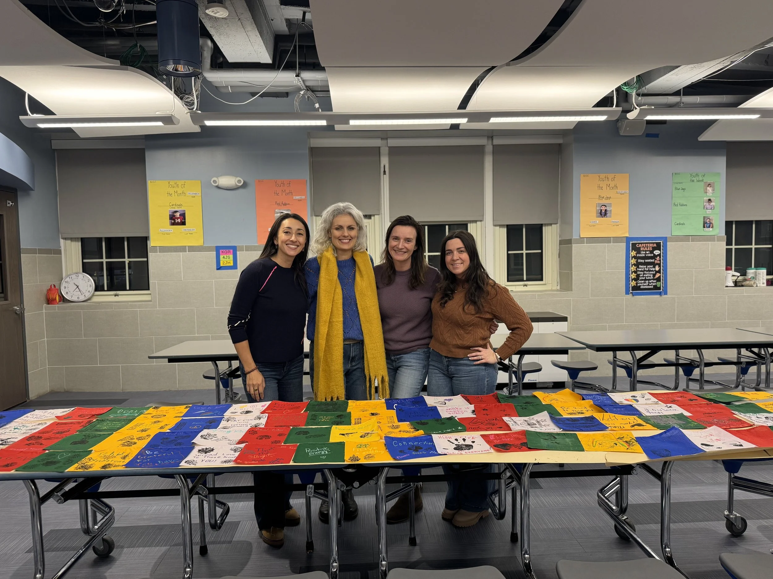 Four women standing behind a table covered with colorful paper crafts in a classroom or school setting.